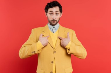 Surprised and stylish man pointing with fingers at himself on coral red background  clipart
