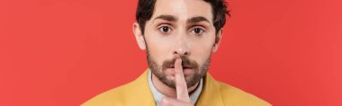 man in yellow blazer holding finger near lips while showing hush sign isolated on red coral background, banner  clipart