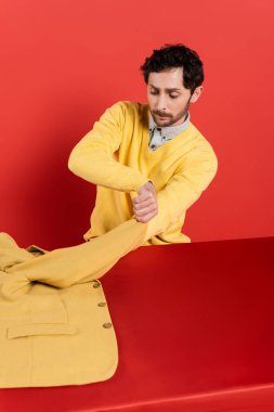 Çekici adam kırmızı mercan arka planda otururken sarı ceketini çıkarıyor. 