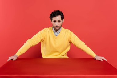 charming guy in yellow long sleeve jumper sitting at table with red tablecloth on coral background  clipart
