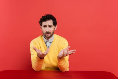 confused man in yellow long sleeve jumper sitting at table and gesturing on coral background  clipart