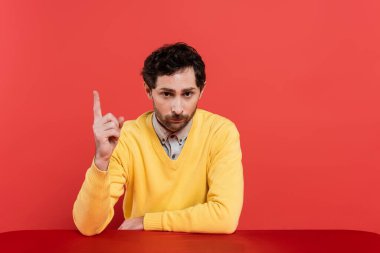 Sarı kollu uzun kollu sakallı adam mercan arka planda tek başına otururken parmağıyla işaret ediyor. 