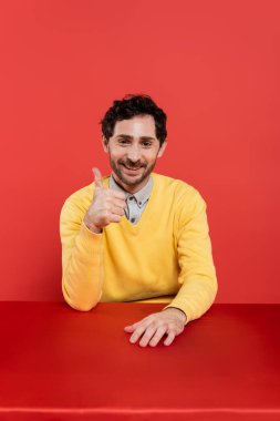 Sarı uzun kollu kazaklı mutlu adam kırmızı mercan arka planda baş parmağını gösteriyor. 