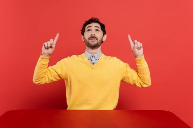 happy man in yellow long sleeve jumper pointing with fingers and looking up on red coral background  clipart