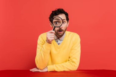scrupulous man in yellow long sleeve jumper holding magnifying glass on red coral background  clipart