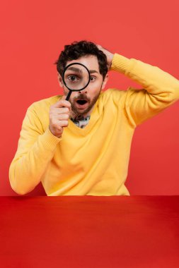 shocked man in yellow long sleeve jumper holding magnifying glass on red coral background  clipart