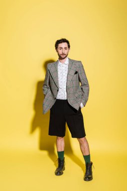 full length of charming guy in grey checkered blazer and shorts standing with hands in pockets on yellow background  clipart