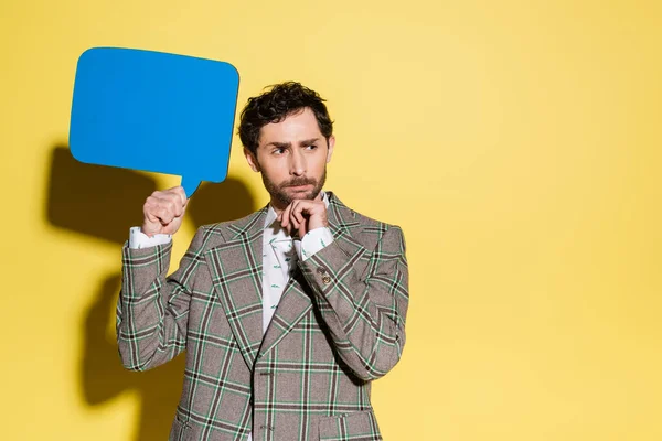 Pensive man in checkered blazer holding speech bubble on yellow background