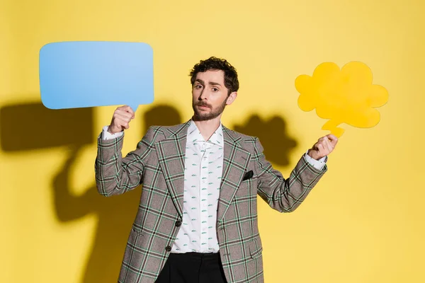 Stylish man holding speech and thought bubbles on yellow background