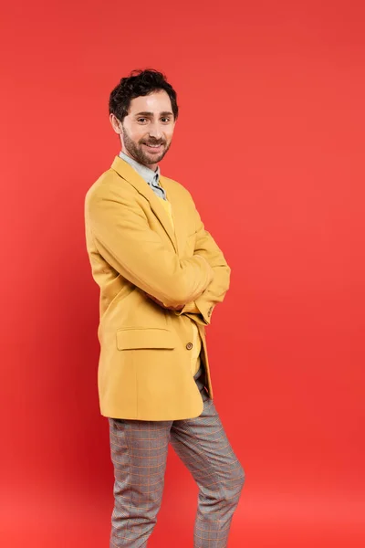 Carefree and stylish man crossing arms and looking at camera isolated on red