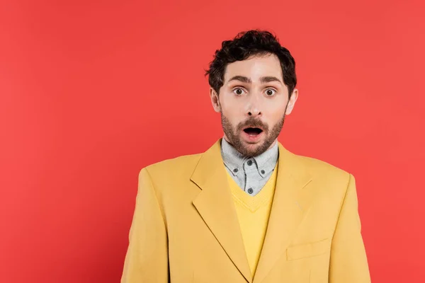 shocked man in yellow blazer standing with opened mouth and looking at camera on coral background