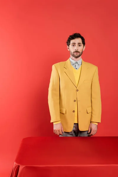 卷曲男子穿着时髦的黄色夹克 站在红色桌子旁边 看着珊瑚背景上的相机 — 图库照片
