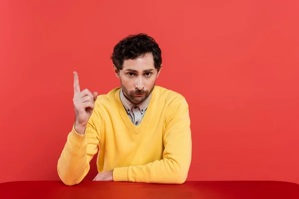 stock image bearded man in yellow long sleeve jumper pointing with finger while sitting isolated on coral background 