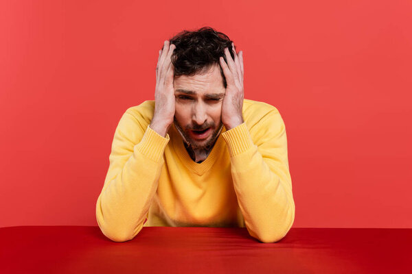 bearded man in yellow long sleeve jumper touching head while having hangover isolated on red coral background 