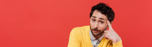 skeptical man in yellow long sleeve jumper with finger near temple looking at camera isolated on coral, banner 