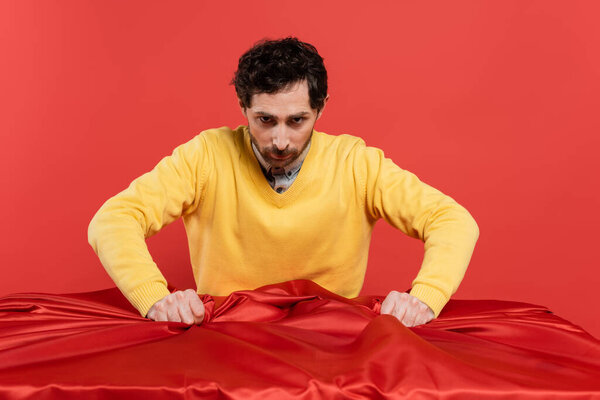 напряженный человек в желтом свитере тянет красную скатерть на стол изолированы на коралловом фоне 