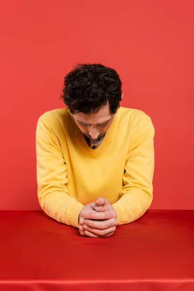 Stock image man in yellow long sleeve jumper sitting with bowed head at table isolated on coral background 