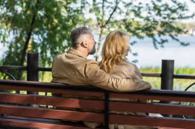 Orta yaşlı bir adam, bahar parkında bankta otururken sarışın karısına sarılıyor. 