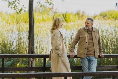 Smiling mature man looking at blonde wife in trench coat on bridge in park  clipart