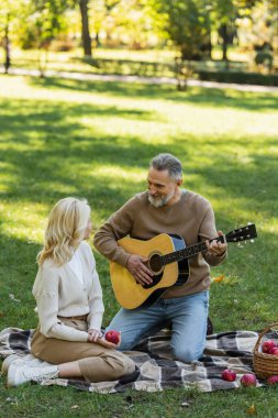 Gri sakallı neşeli orta yaşlı bir adam parkta piknik yaparken sarışın karısının yanında akustik gitar çalıyor. 