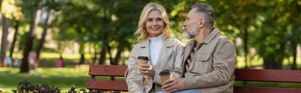笑顔ブロンド女性保持テイクアウトコーヒー近く夫上のベンチで春の公園 バナー — ストック写真