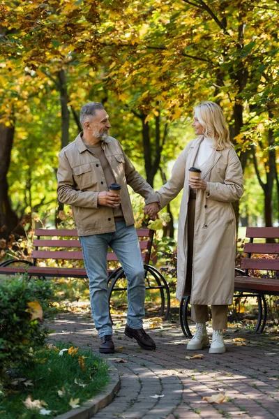 公園を歩いている間に行くために手とコーヒーを握っている成熟したカップルの笑顔の側面図 — ストック写真