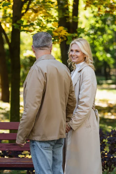 在模糊的公园里微笑的金发女人牵着丈夫的手 — 图库照片