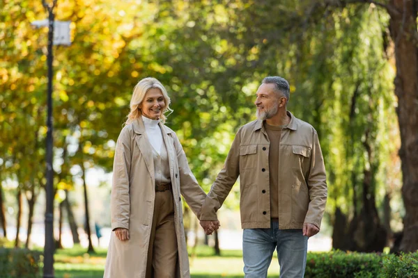Happy mature man holding hand of blonde wife in trench coat in park