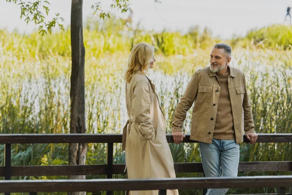 笑着成熟的男人看着金发碧眼的妻子穿着风衣在公园的桥上 — 图库照片