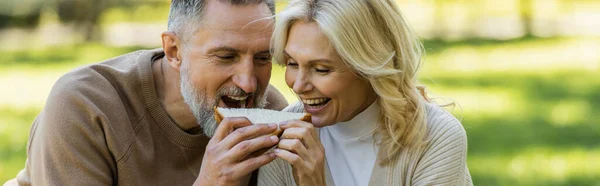 陽気な中年のカップルが両側からサンドイッチを噛んだ — ストック写真
