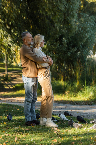 full length of happy middle aged man hugging blonde wife while standing near pigeons in park 