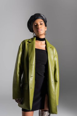 Young african american woman in leather beret and coat looking at camera isolated on grey  clipart