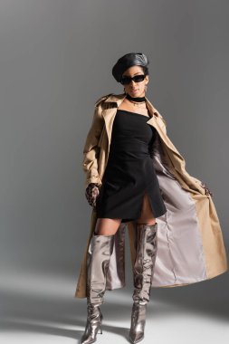 Trençkot ve gri arka planda parlak botlarla poz veren modaya uygun Afrikalı Amerikalı kadın.