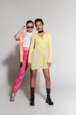 Güneş gözlüklü modaya uyan Asyalı kadın, gri arka planda Afrikalı Amerikalı arkadaşının yanında poz veriyor. 