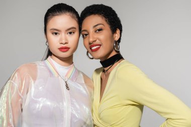 Gri yolda yalnız kalmış Asyalı arkadaşının yanında gülümseyen modaya uyan Afrikalı Amerikalı kadın.  