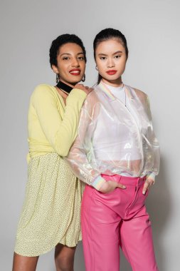 Gülümseyen Afrikalı Amerikalı kadın gri arka plandaki Asyalı arkadaşının yanındaki kameraya bakıyor. 