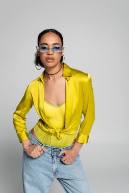 Moda mavisi güneş gözlüklü şık Afro-Amerikalı kadın gri üzerine izole edilmiş poz veriyor. 