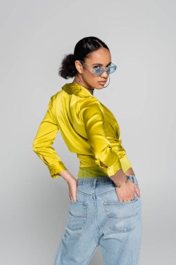 young african american model in blue sunglasses and trendy clothes posing with hands in back pockets isolated on grey  clipart