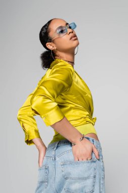 Mavi güneş gözlüklü ve şık elbiseli Afro-Amerikan modeli. Gri cepli ellerle poz veriyor. 