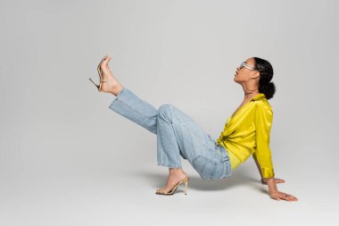 Yüksek topuklu ve gri arka planda poz veren modaya uygun güneş gözlüklü Afro-Amerikan kadının yan görüntüsü 