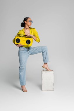 full length of brunette african american model stepping on concrete cube while holding yellow boombox on grey  clipart