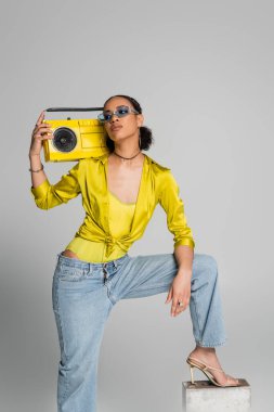 Esmer, Afro-Amerikalı, mavi güneş gözlüklü, beton küpün üstüne basan, elinde sarı müzik seti olan gri gözlüklü kadın. 