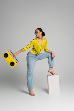 full length of brunette african american woman in sunglasses stepping on concrete cube while holding retro boombox on grey  clipart