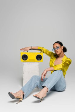 full length of brunette african american woman in sunglasses sitting near concrete cube and retro boombox on grey  clipart