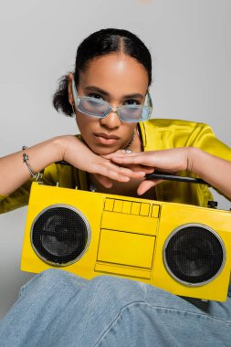 Mavi güneş gözlüklü şık Afrikalı Amerikalı kadın. Gri renkte izole edilmiş sarı retro müzik kutusu yanında poz veriyor. 