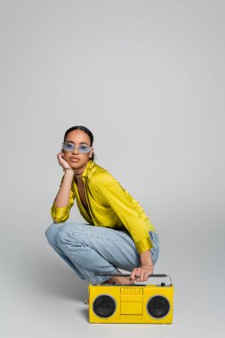 full length of brunette african american woman in blue sunglasses sitting on haunches near yellow boombox on grey  clipart