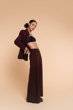 full length of slim african american model in crop top and stylish maroon suit with wide leg pants posing on beige  clipart