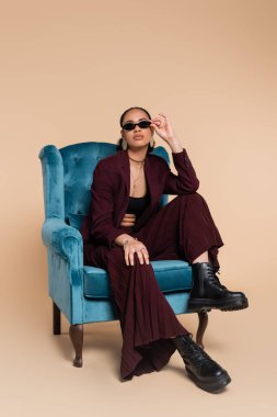 Şık Afro-Amerikan modeli kestane rengi takım elbiseli ve son moda güneş gözlüklü bej renkli mavi kadife koltukta oturuyor. 