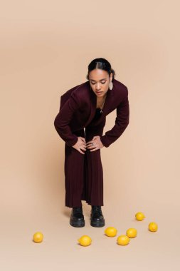 stylish african american woman in burgundy suit standing and looking at fresh lemons on beige  clipart