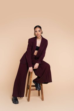 full length of stylish african american woman in burgundy suit sitting on wooden high stool on beige  clipart